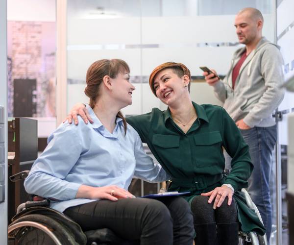 Two women in wheelchairs smile at each other, one provides NDIS Complex Care