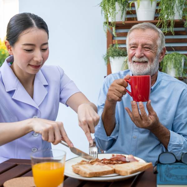 A female NDIS Personal Care worker cuts breakfast food for a smiling elderly man