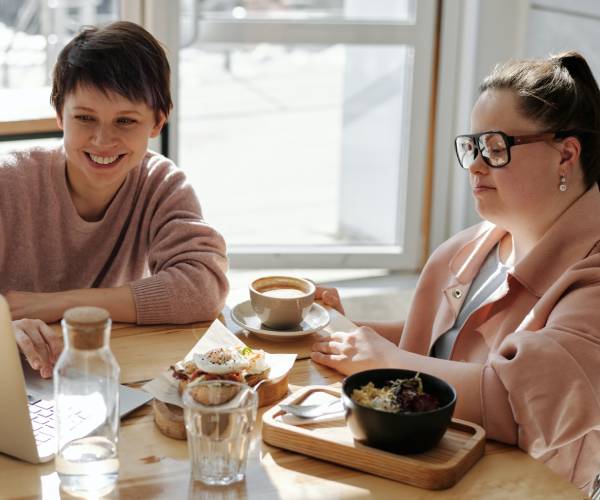 A young man with Down Syndrome smiles at his NDIS Supported Independent Living support worker while on a laptop