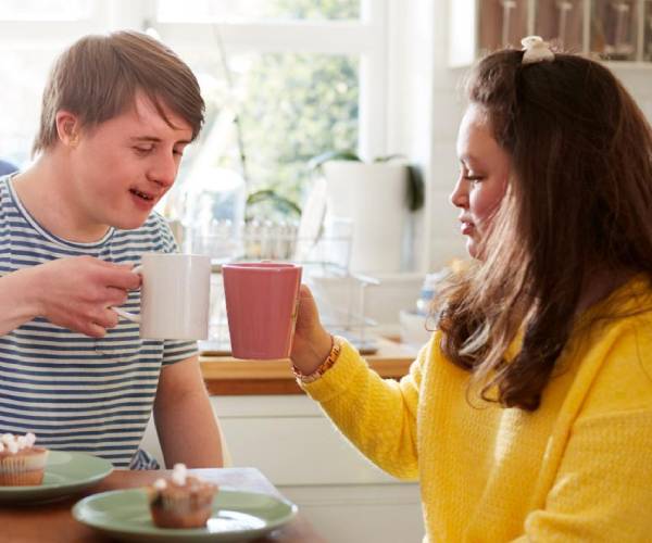 A carer helps an older woman with a cane stand up indoors